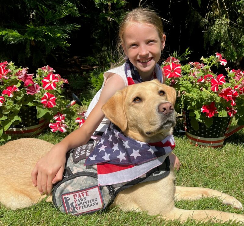 Yellow Lab Nutmeg wearing her service vest and American flag bandana posing with a young girl in front of potted flowers.