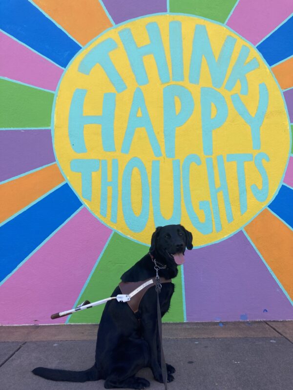 Tawny sits in front of a colorful mural that says ‘Think Happy Thoughts’
