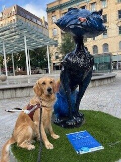 <p>'Moon', a petite golden retriever, sits in harness next to a 'Coraline' cat statue in downtown Portland. The cat statue is about twice her height and is painted to look like the night sky. </p>