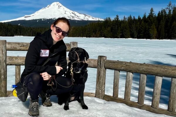 Black Lab Cinder and her trainer posing in a snowy/icy area with a perfect view of Mt. Hood in the background.